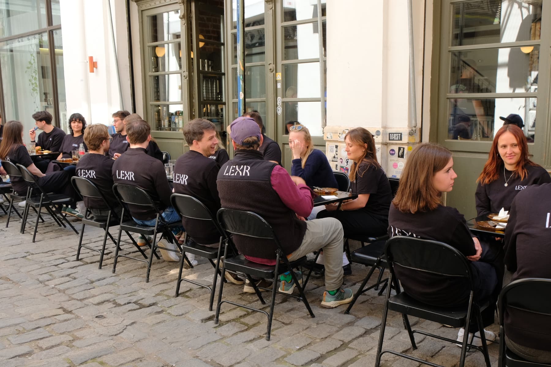 Colleagues eating lunch on black folding chairs along a shopfront on a cobbled street