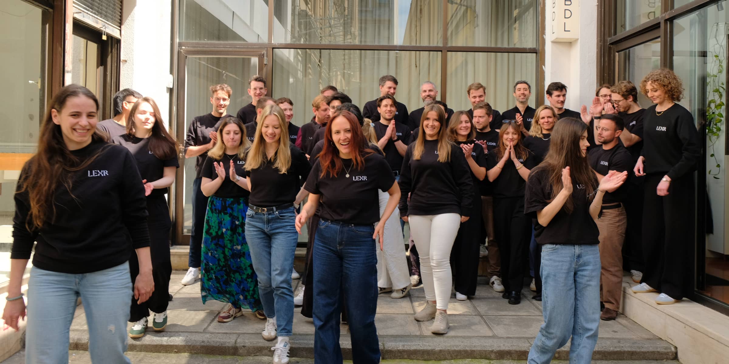 About thirty LEXR colleagues in black LEXR shirts walking out of a glass-fronted courtyard, several of them applauding