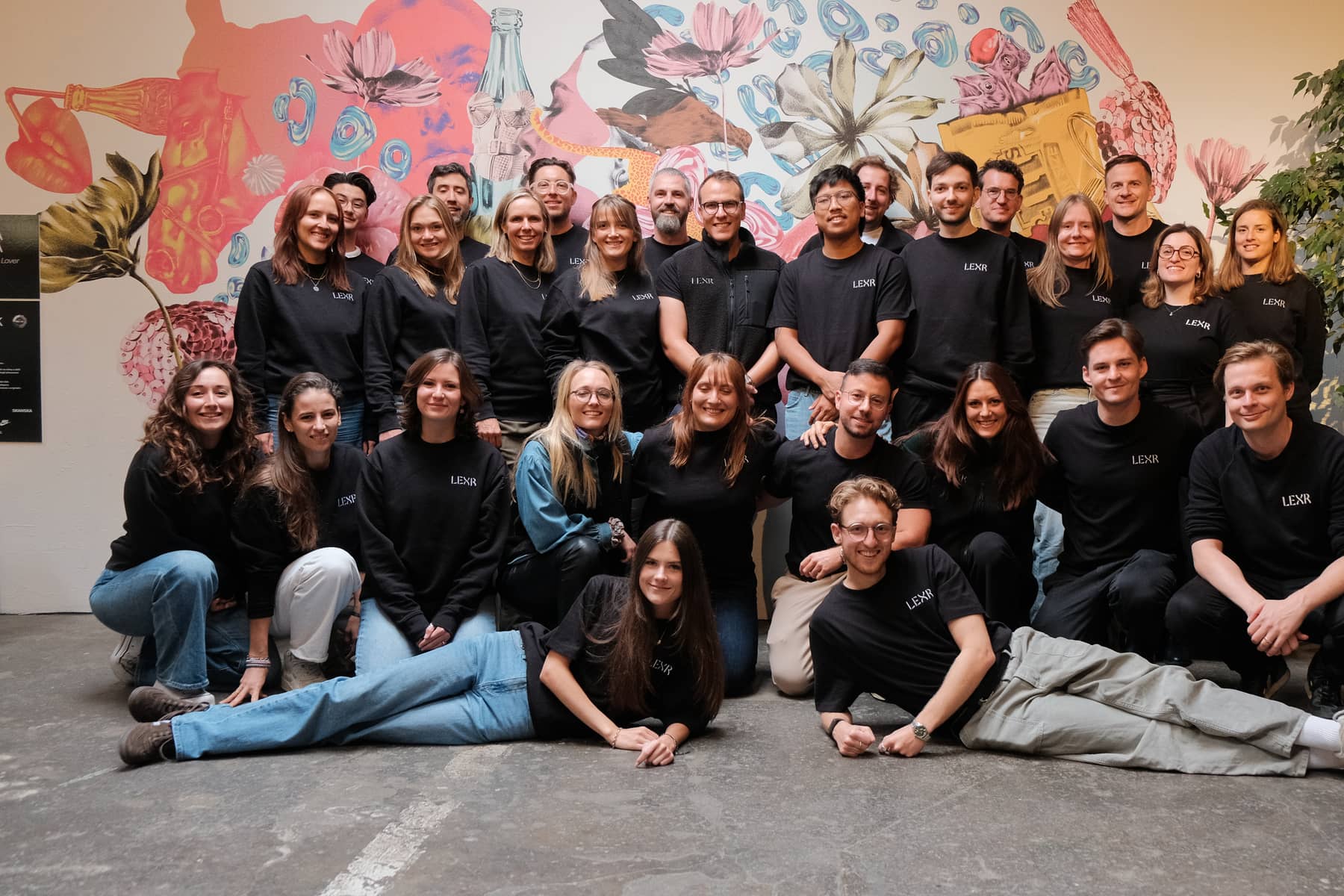 The LEXR team in three rows in front of a large floral mural, two people lying on the floor at the front