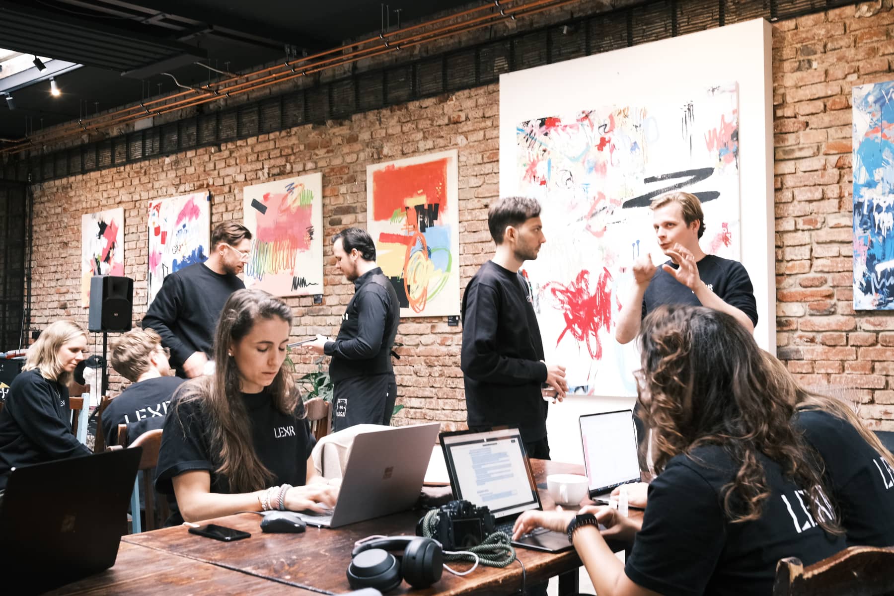 Colleagues working at a long table in a brick hall hung with large abstract paintings, two of them standing and talking